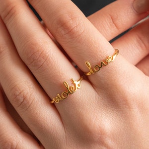 May include: Two gold rings with the words "love" and "violet" on them. The rings are worn on the ring finger of a person's hand.