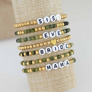 Puede incluir: Conjunto de pulseras de cuentas con letras blancas que deletrean "SISTERS", "EVE", "BRIDE" y "MAMA". Las pulseras presentan cuentas doradas y verdes, con un dije de corazón dorado.