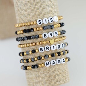 May include: A collection of beaded bracelets with white letter beads spelling out "SIS", "EVE", "BRIDE", and "MAMA". The bracelets feature gold and black beads, and a gold heart charm. The bracelets are displayed on a tan fabric.
