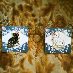 May include: Two enamel pins featuring a rat design. One pin is black with a crescent moon and the other is white. Both rats wear crowns and hold a star. The pins are on cards with a floral pattern.