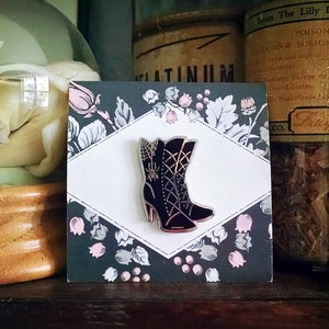 May include: Black enamel pin featuring a pair of black boots with a spider web design and a spider on the front. The pin is on a card with a floral pattern and the word "PLATINUM" printed on it.