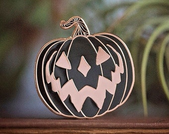May include: Black and gold enamel pin of a jack-o'-lantern with a spooky expression. The pumpkin is outlined in gold and has a detailed face with a wide grin and diamond-shaped eyes.