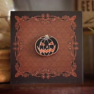 May include: A black and orange Halloween pumpkin enamel pin with a gold border. The pin is set on a brown card with an ornate orange border. The pumpkin has a carved face design. A festive accessory for Halloween.