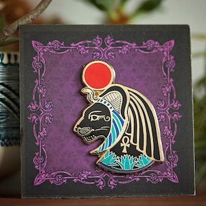 May include: Gold-toned enamel pin depicting a black cat's head with a red sun, blue and white headdress, and an ankh symbol. The pin is displayed against a purple background with a decorative gold frame.