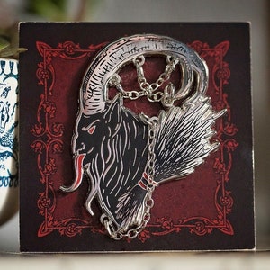 May include: Silver-toned enamel pin featuring a black and white illustration of a goat's head with horns, a long tongue, and a chain around its neck. The background is a dark red with a decorative border.