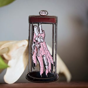 May include: A silver-toned enamel pin depicting a pink skeletal hand inside a glass jar with a diamond-shaped lid. The hand is partially submerged in a pink liquid.