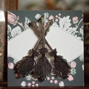 May include: A silver brooch depicting three crossed witch brooms. The brooch is set against a dark grey card with a floral pattern in pink and white. The design is a decorative accessory, suitable for clothing or bags.