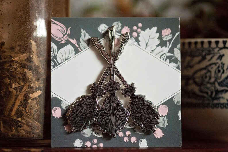 May include: A silver brooch depicting three crossed witch brooms. The brooch is set against a dark grey card with a floral pattern in pink and white. The design is a decorative accessory, suitable for clothing or bags.