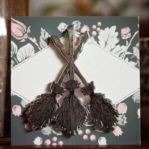 May include: A silver-coloured pin depicting three crossed brooms. The brooms have dark grey bristles and are set against a grey background with a floral pattern and a white diamond-shaped space.
