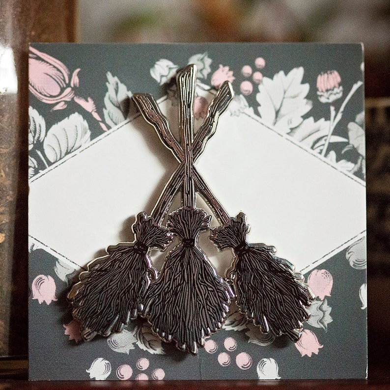 May include: A silver-coloured pin depicting three crossed brooms. The brooms have dark grey bristles and are set against a grey background with a floral pattern and a white diamond-shaped space.