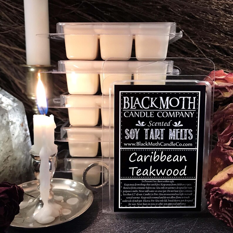 May include: Soy tart melts in clear packaging, labelled "Black Moth Candle Company" with "Caribbean Teakwood" scent. A lit candle and other candles are in the background, creating a warm ambiance.