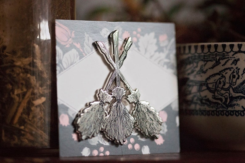 May include: A silver brooch depicting three stylized brooms, set on a grey card with a floral pattern and a white diamond-shaped area. The brooms have detailed designs, possibly related to craft or fantasy.