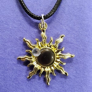 May include: A gold sun pendant necklace with a black cord. The sun has a dark gray center and a clear rhinestone accent.