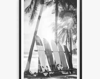 Puede incluir: Impresión en blanco y negro enmarcada con tablas de surf apoyadas en un estante de madera en una playa. Se ven palmeras y luz solar en el fondo, creando una escena tropical. La imagen es una fotografía en escala de grises.
