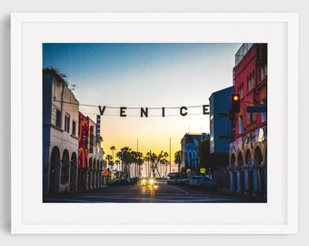 May include: A street scene in Venice, California, with a sign that reads "VENICE" in black letters. The street is lined with colorful buildings and palm trees. The sky is a vibrant blue with a hint of orange from the setting sun.