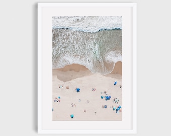 May include: An aerial view of a beach with white sand and blue water. The waves are crashing on the shore and there are people swimming and sunbathing. There are also blue umbrellas scattered across the beach.