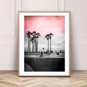 May include: Black and white photograph of a skateboarder performing a trick in a skate park. The background features palm trees and a pink sunset.