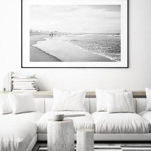 May include: Black and white photograph of a beach scene with people walking along the shore. The photo is framed in a black frame and hanging on a white wall above a white couch.