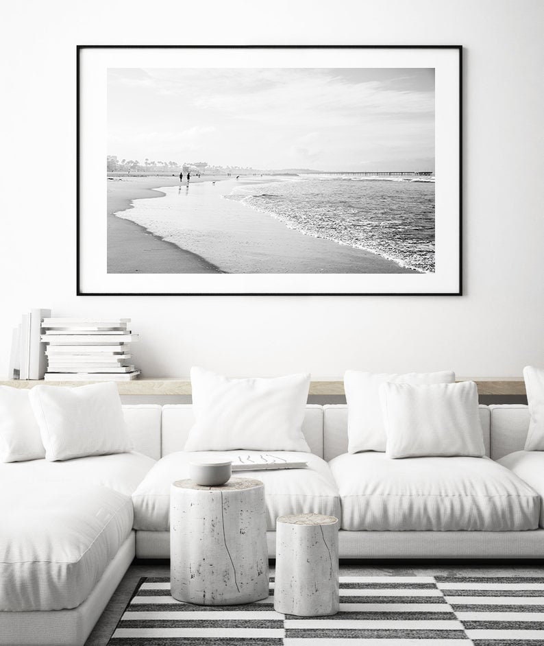May include: Black and white photograph of a beach scene with people walking along the shore. The photo is framed in a black frame and hanging on a white wall above a white couch.