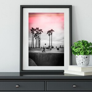 May include: Black and white photograph of a skateboarder in a skate park with palm trees in the background. The sky is a vibrant pink.
