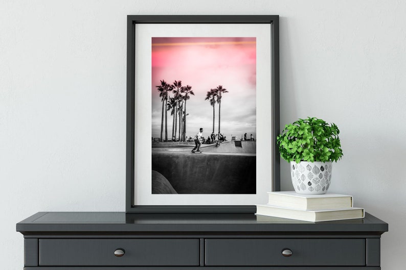 May include: Black and white photograph of a skateboarder in a skate park with palm trees in the background. The sky is a vibrant pink.