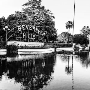 May include: A black and white photo of a sign that reads "Beverly Hills" with a pond in the foreground and palm trees in the background.