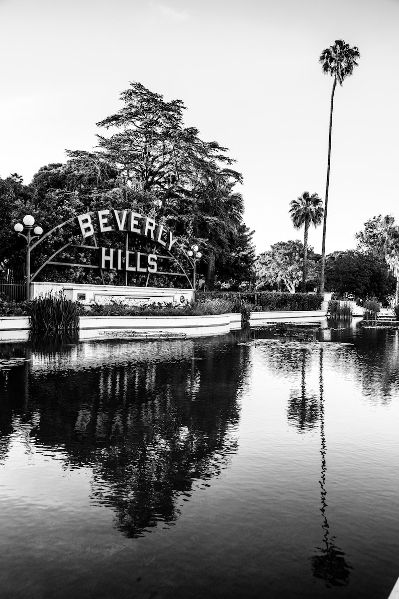 May include: A black and white photo of a sign that reads "Beverly Hills" with a pond in the foreground and palm trees in the background.