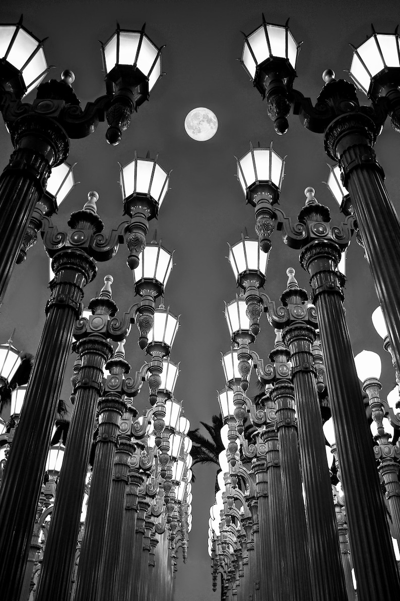 May include: A black and white photo of a row of street lamps with a full moon in the background. The lamps are tall and slender with ornate designs. The moon is bright and round.