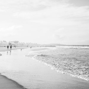 May include: A black and white photograph of a beach with a pier in the distance. The beach is empty except for a few people walking along the shore.