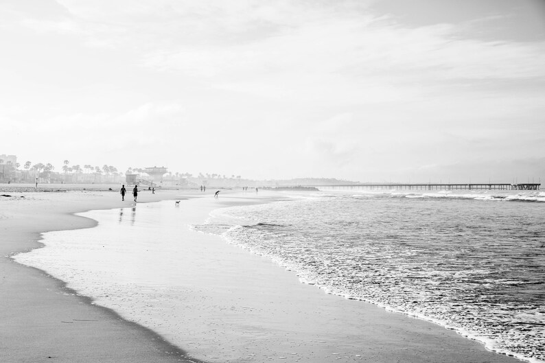 May include: A black and white photograph of a beach with a pier in the distance. The beach is empty except for a few people walking along the shore.
