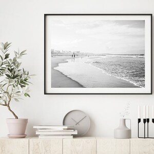 May include: Black and white photograph of a beach scene with people walking along the shore. The photo is framed in a black frame and hanging on a white wall.