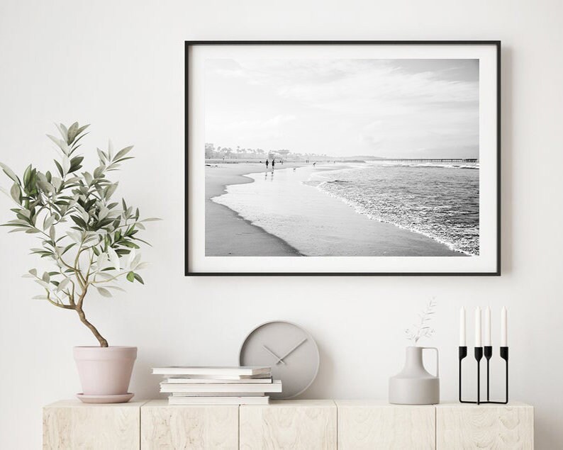 May include: Black and white photograph of a beach scene with people walking along the shore. The photo is framed in a black frame and hanging on a white wall.