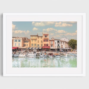 Op de afbeelding: Een ingelijste prent van een kustlandschap met kleurrijke gebouwen, boten en een rustige haven. De gebouwen zijn geschilderd in tinten geel, blauw en rood. De lucht is lichtblauw met witte wolken. Verschillende boten liggen in het water.