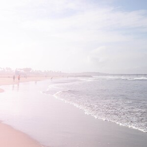 May include: A hazy, pink-hued beach scene with a wooden pier in the distance. The sand is white and the water is a light blue. There are a few people walking on the beach.