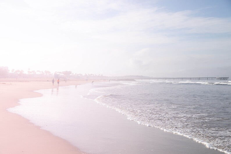 May include: A hazy, pink-hued beach scene with a wooden pier in the distance. The sand is white and the water is a light blue. There are a few people walking on the beach.