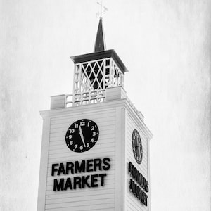 May include: A black and white photo of a white building with a clock tower. The clock tower has a clock face with black hands and numbers. The building has a sign that says "Farmers Market".