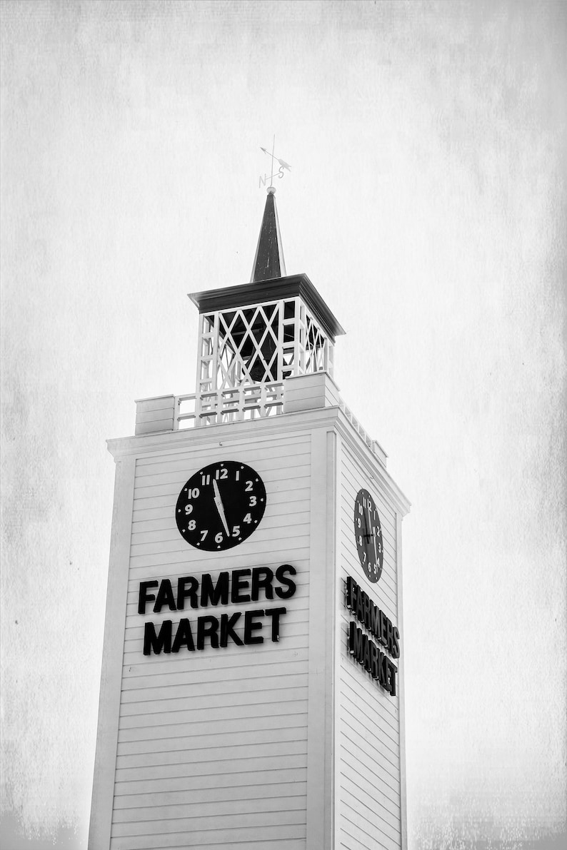 May include: A black and white photo of a white building with a clock tower. The clock tower has a clock face with black hands and numbers. The building has a sign that says "Farmers Market".