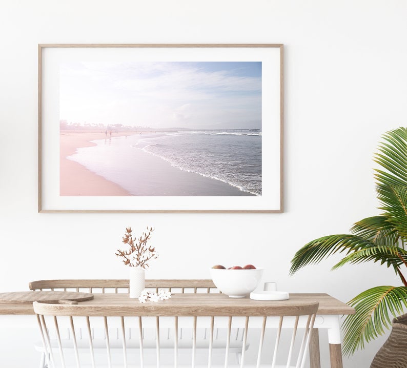 May include: A framed photograph of a beach scene with a soft pink and blue color palette. The photo shows a sandy beach with waves crashing on the shore. The sky is a light blue with white clouds.