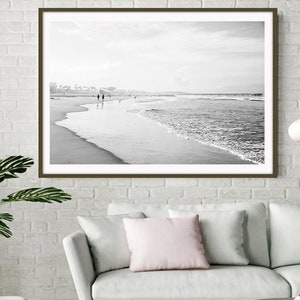 May include: Black and white photograph of a beach scene with a couple walking along the shore. The ocean waves are crashing on the sandy beach. The photo is framed in a brown frame.