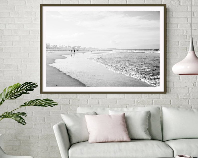 May include: Black and white photograph of a beach scene with a couple walking along the shore. The ocean waves are crashing on the sandy beach. The photo is framed in a brown frame.