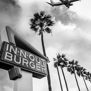 May include: A black and white photo of a In-N-Out Burger sign with a plane flying overhead. The sign is in the shape of a triangle and has the words "In-N-Out Burger" written on it. There are palm trees in the background.