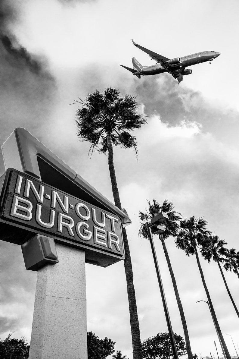 May include: A black and white photo of a In-N-Out Burger sign with a plane flying overhead. The sign is in the shape of a triangle and has the words "In-N-Out Burger" written on it. There are palm trees in the background.