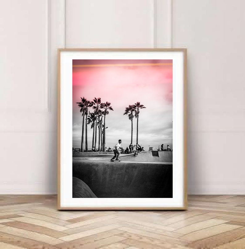May include: Black and white photograph of a skateboarder performing a trick in a skate park. The background features palm trees and a pink sunset.