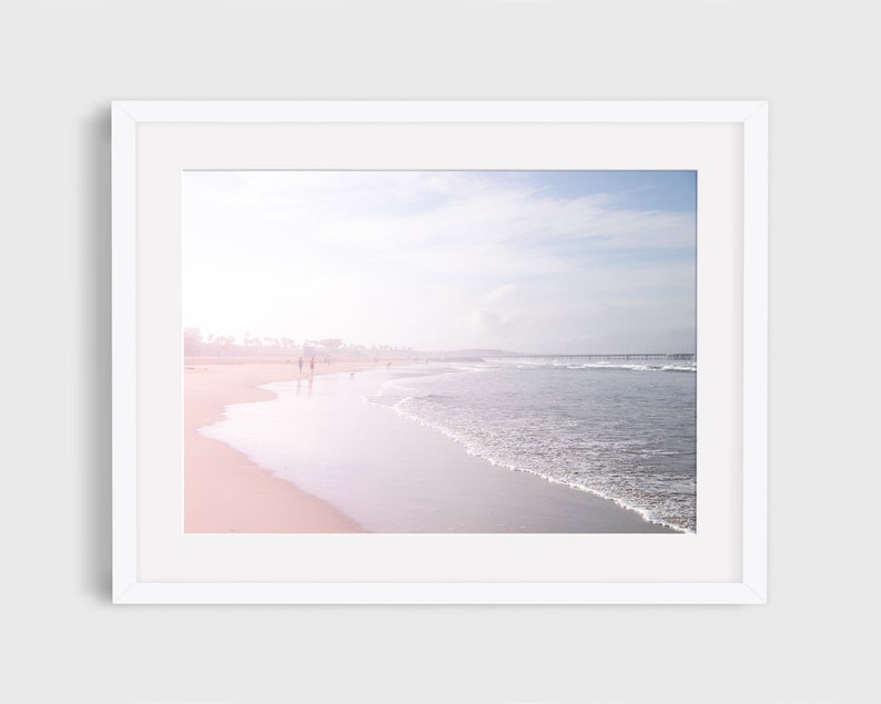 May include: A photograph of a beach scene with a soft, pink and blue color palette. The photo shows a sandy beach with waves crashing on the shore. There are people walking along the beach in the distance.