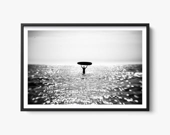 Può includere: Fotografia in bianco e nero di una persona in piedi nell'oceano che tiene una tavola da surf sopra la testa. L'acqua brilla e il cielo è sereno.