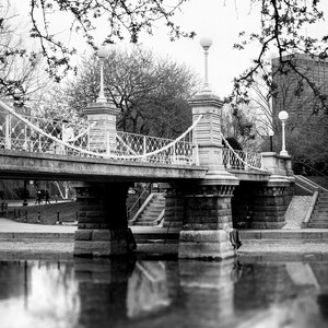 Puede incluir: Una foto en blanco y negro de un puente de piedra con una barandilla de hierro forjado. El puente est&aacute; sobre una masa de agua y hay &aacute;rboles y edificios en el fondo.