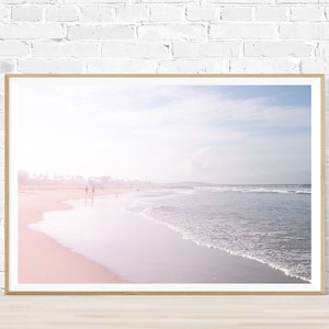 May include: A framed photograph of a beach scene with a soft, pink and blue color palette. The photo shows a sandy beach with waves lapping at the shore. There are a few people walking in the distance.