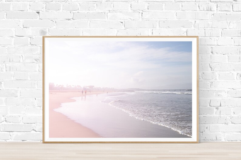 May include: A framed photograph of a beach scene with a soft, pink and blue color palette. The photo shows a sandy beach with waves lapping at the shore. There are a few people walking in the distance.