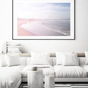 May include: A framed photograph of a beach scene with a soft, pink and blue color palette. The photo shows a sandy beach with waves crashing on the shore. There are people walking along the beach in the distance.