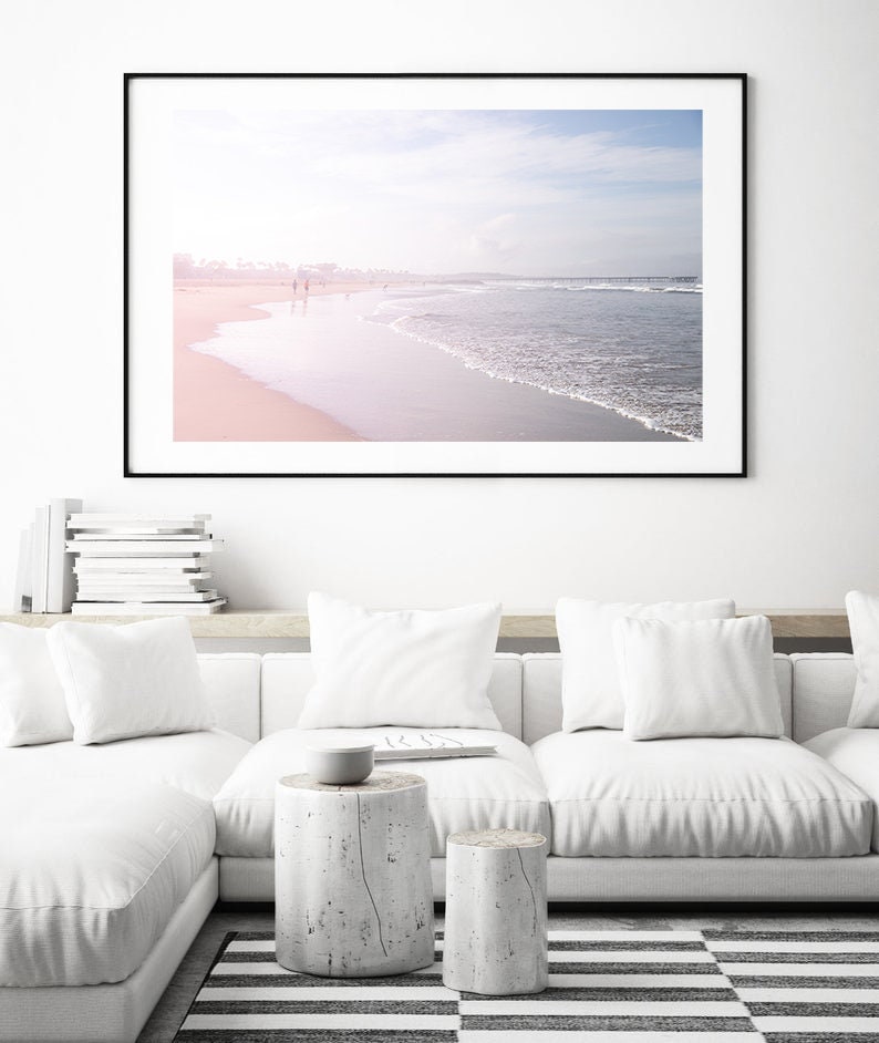 May include: A framed photograph of a beach scene with a soft, pink and blue color palette. The photo shows a sandy beach with waves crashing on the shore. There are people walking along the beach in the distance.